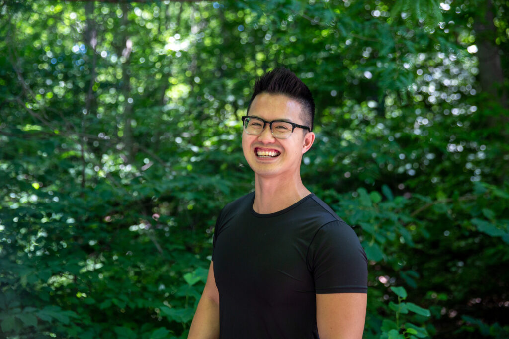 Joshua Nguyen, wearing a black shirt and glasses, smiles at the camera while standing in front of green foliage.