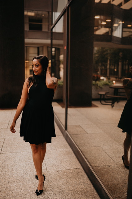 Rita Mookerjee stands in front of a big glass building. She is a brown femme with long dark hair wearing a sleeveless little black dress. She is looking to the side with one hand in her hair.
