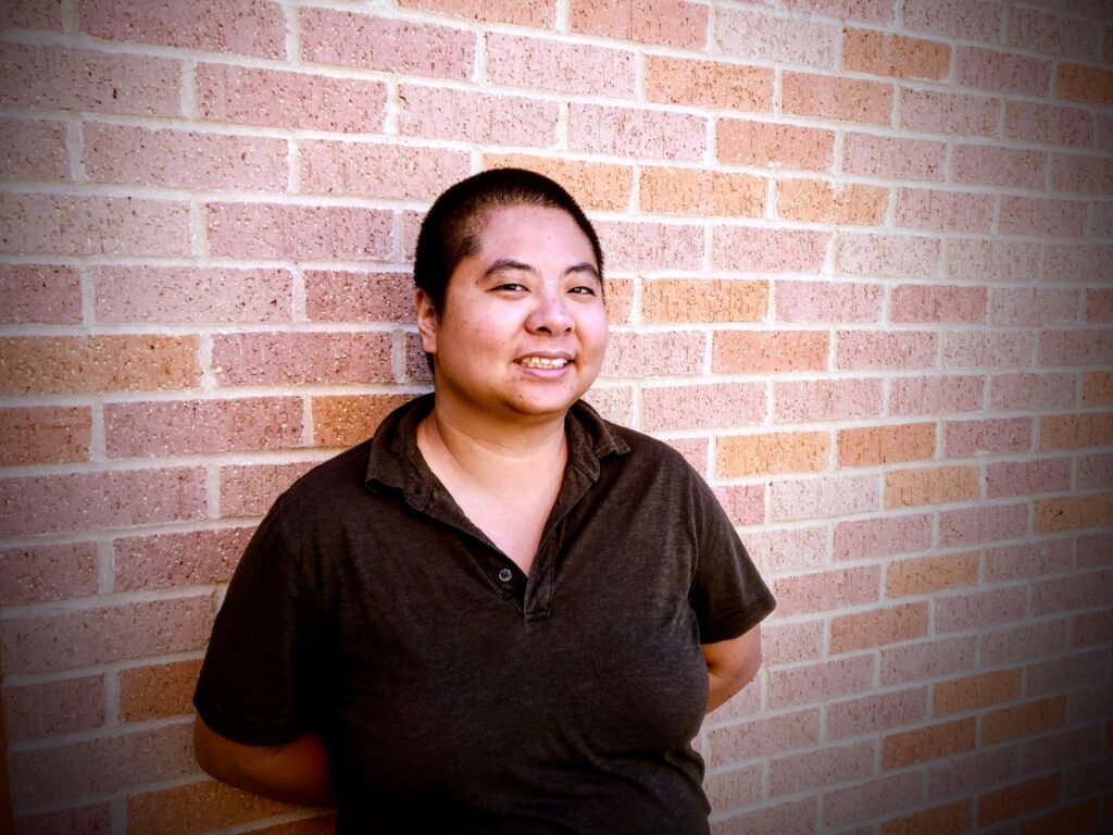 An Asian American person wearing a dark shirt stands in front of a brick wall and smiles.