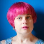 Jill Khoury has short pink hair and pale skin with pink lipstick. In front of a blue background, she gazes thoughtfully at the camera.