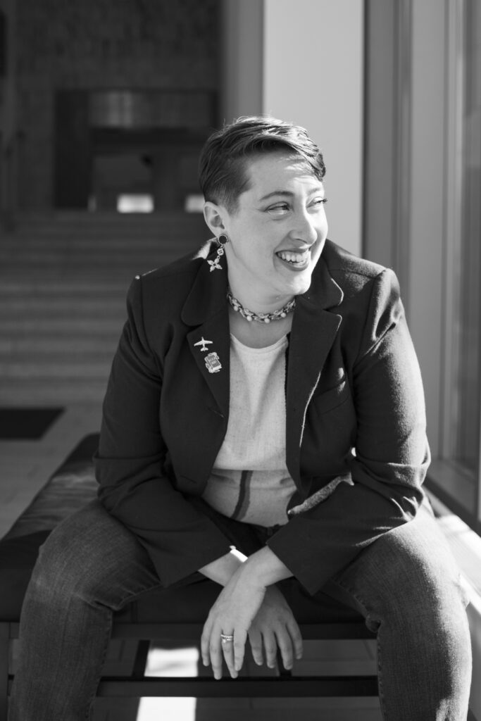 A black and white photo of Mandy Shunnarah, a writer who uses they/them pronouns. Mandy has short brown hair and pale skin. They are wearing a blazer and sitting leaned over with their elbows on their knees. Their face is turned to the side, and they are smiling as the sun shines on their face.