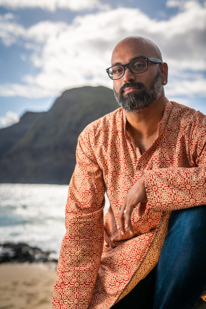 A Guyanese man with a salt-and-pepper beard kneels on the beach, in front of a green mountain. He wears a long-sleeve, red and light-orange patterned shirt, and blue jeans. He has thick black framed glasses, a silver nose ring, and silver rings on his left hand.