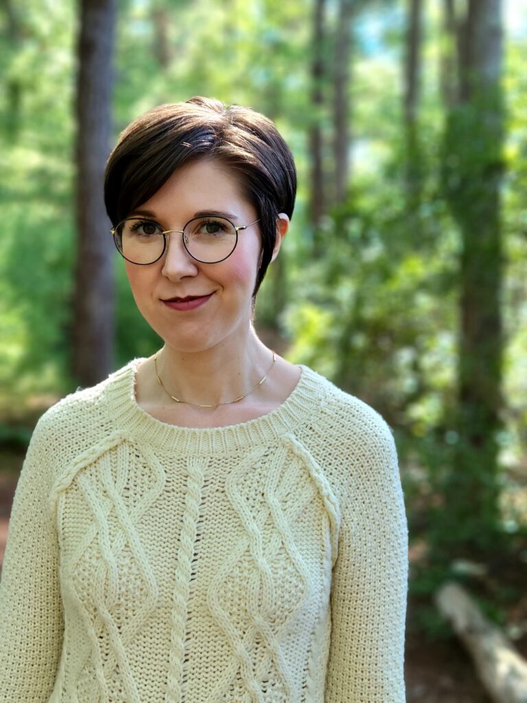 A pixie haired woman wearing round glasses and a cream-colored cable knit sweater smiles at the camera while standing in a bright forest backdropped by green trees and blue sky.