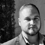 Black-and-white headshot of Clayton Bradshaw-Mittal with a short beard and short hair from the shoulders up smiling in a suit with no tie.