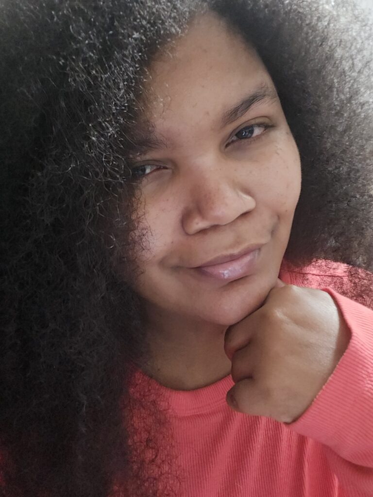 Portrait-oriented selfie of Ashley Elizabeth smirking and wearing a coral pink shirt in 3/4th profile, her long, coiled, dark brown hair over a shoulder and taking up the majority of the background, Her chin rests on her hand.