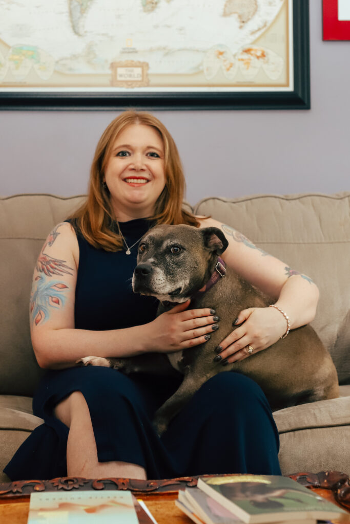 Tattooed middle-aged white woman with strawberry blonde hair and fair skin smiling while wearing a navy blue sleeveless dress, holding a tubby grey pitbull, and sitting on a couch.