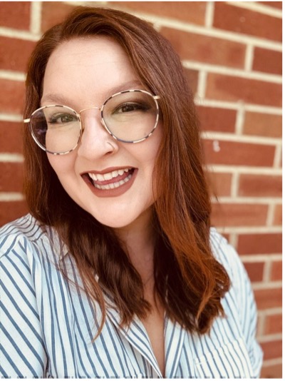 Image is a white woman with long red hair and glasses. She stands in front of a brick wall and wears a striped white and grey shirt. She's smiling.
