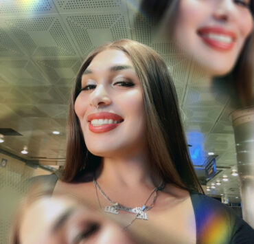 a light skinned Mexican Indigenous woman smiles brightly at the viewer. She has slick brown hair parted in the middle and wears and silver hoops, septum, and necklaces that read: “310”, “iká”, and “shumarí”. She wears a black open chested blouse with long sleeves. The background has colorful light streaks.