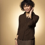 A professional headshot of Franchesca Nicole Lazaro, a Filipino-American woman with short curly dark hair and bangs, smiling at the camera. She wears a dark brown button-up blouse and a brown skirt, accessorized with beaded bracelets and earrings. Her left hand is raised near her face in a relaxed pose. The photo is taken against a warm, brown-beige gradient background