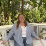 A white woman with blonde hair and clear glasses sitting on a couch outside with several trees and plants in the background. She is wearing jeans and a blue-and-white stripped button down shirt.