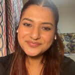 A South-Asian woman smiling at the camera, with a Doctor Who poster visible in the background. She has long, dark auburn hair and is wearing a black shirt.