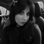 Picture of a young woman with bangs and long dark hair which covers her shoulders and the necklace she is wearing, leaving only the chain showing. There is a bedazzled bow-shaped hair clip on the left side of the image. Her eye makeup and lipstick appears rather dark, contrasting her fair complexion. She is in a car, and the photo is in black and white. She looks into the camera with a neutral expression, angling her face to pose for a selfie.