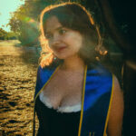 A white woman is standing in front of a tree in a grove. She has short, dark red hair. She is wearing a black dress with white trim and a blue graduation stole with the words "UC Irvine" embroidered on it with gold thread.
