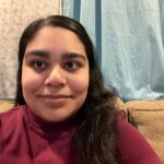 A South Asian woman smiling and sitting on a green couch. She has black hair, half lying over her right shoulder and half down her back, and is wearing a wine-red turtleneck. Behind her are two closed window curtains, one light blue and floral print, and the other solid teal.