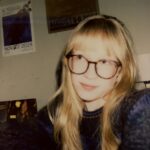A young white woman with long blonde hair, bangs, and circular glasses, barely smiling at the camera, stares up at the corner of the screen, posed against a tan background with music festival posters.