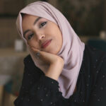 A headshot of a woman wearing a pale pink headscarf and a black blouse with white polka dots, looking at the camera with her palm resting against her face.