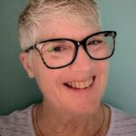 Face of a white woman with short gray hair parted to the side against a teal background. She is smiling and wearing large black glasses, a pale lavender sweater with stripes, and a circular silver necklace with colored beads.