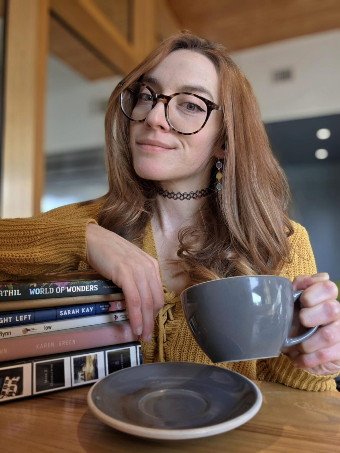 A feminine white person with long red hair and thick glasses smirking while holding a coffee cup in one hand and their arm on a stack of books