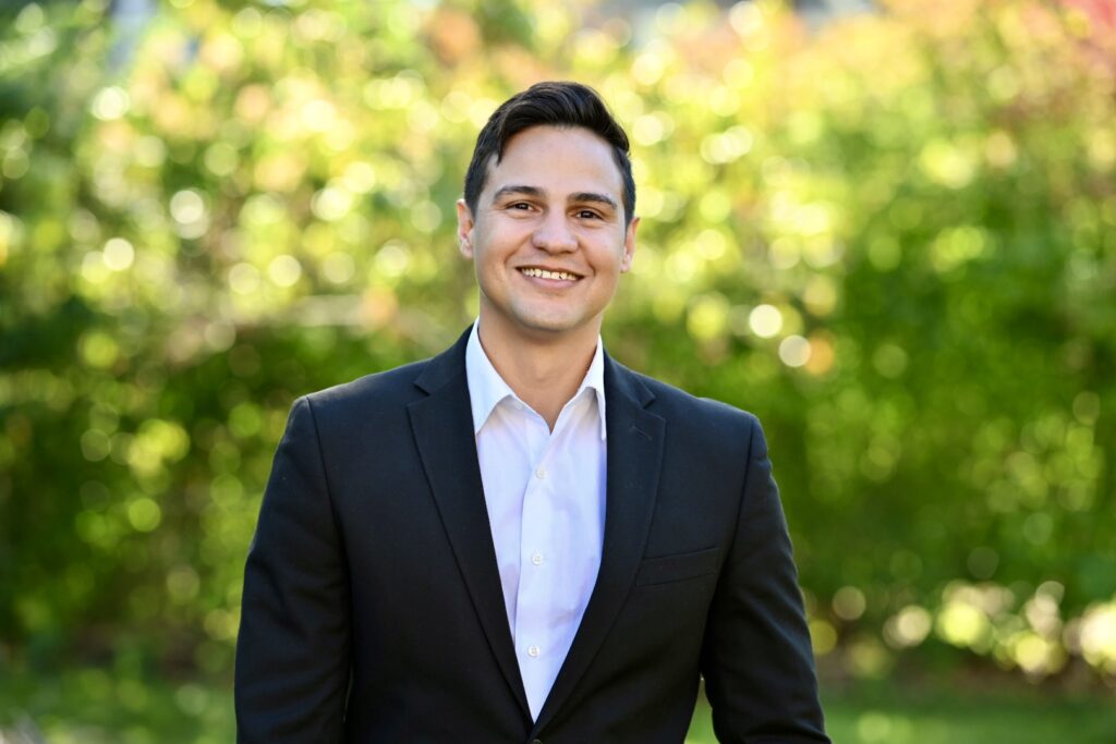 Image shows a smiling person with short dark hair wearing a black blazer over a white button-down shirt, standing outdoors against a blurred background of green foliage and sunlight.