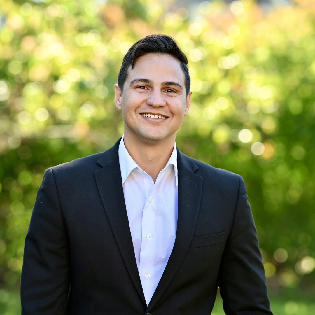 Ayendy Bonifacio - Ríos are for Chivos and Eternity Image shows a smiling person with short dark hair wearing a black blazer over a white button-down shirt, standing outdoors against a blurred background of green foliage and sunlight.