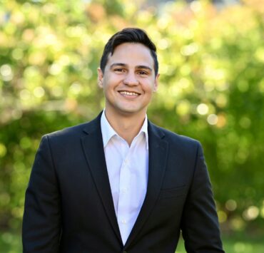 Image shows a smiling person with short dark hair wearing a black blazer over a white button-down shirt, standing outdoors against a blurred background of green foliage and sunlight.