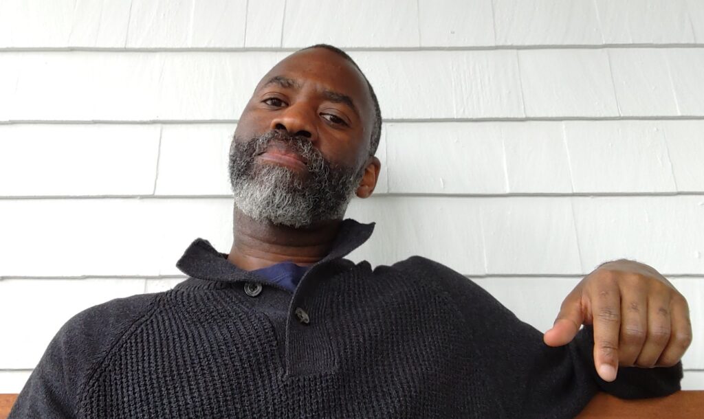A Black man with a greying beard, in a grey sweater, sitting in front of a white wall, looking toward the viewer.
