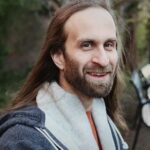 a smiling person with long, brown hair and a beard, wearing a grey hoodie over an orange shirt, poses with an out-of-focus park bench and trees behind them. Photo taken by Jessica Lynne Furtado.