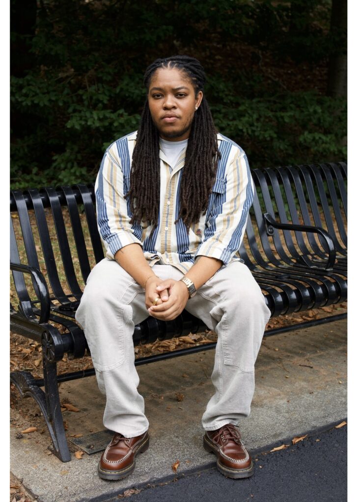 The photo is of a Black transmasculine man with long locs, a tan and blue striped shirt, tan pants, and red-brown loafers. He is sitting on a park bench with his hands clasped in front of him.