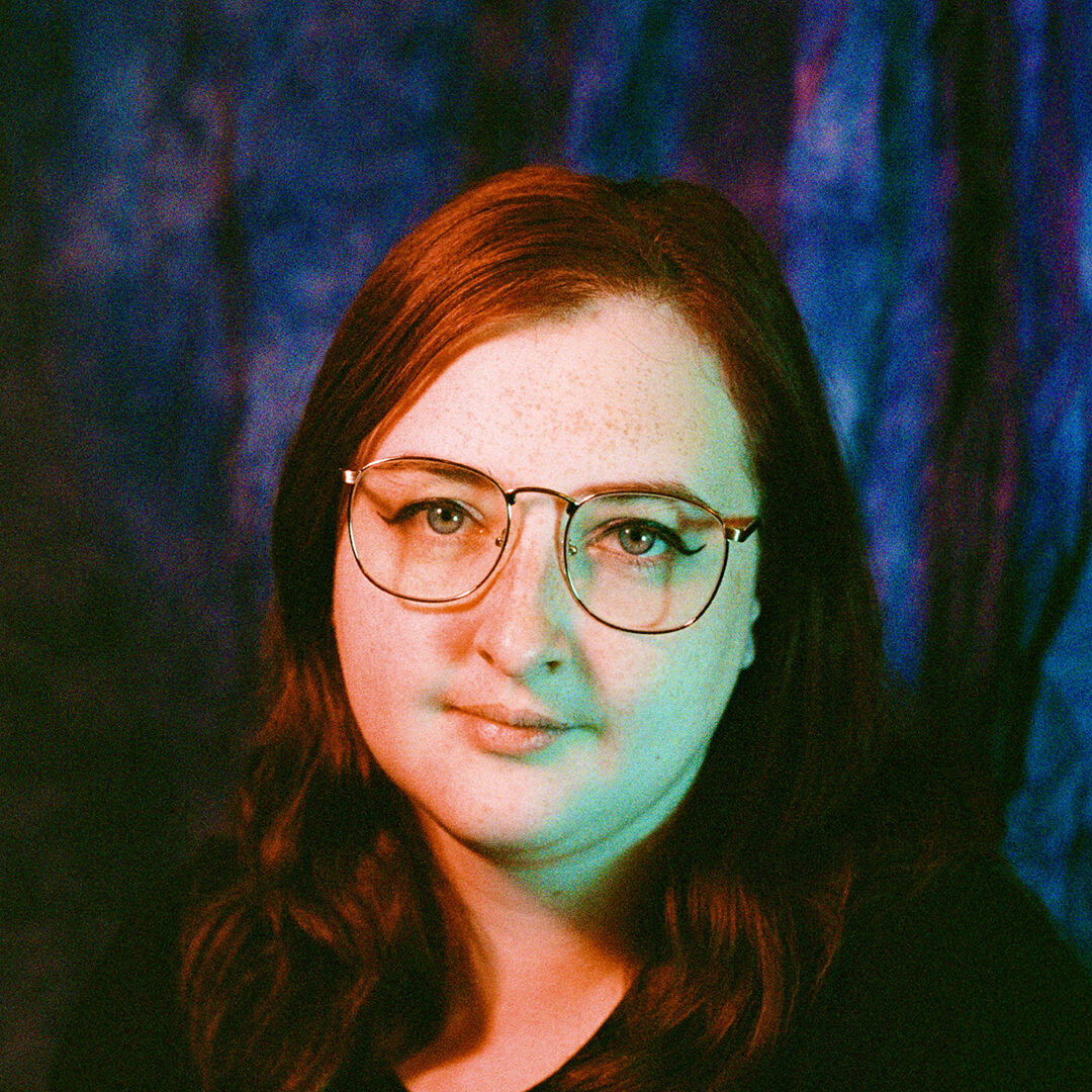A 35mm film portrait of nat raum, a white agender person with medium-length red hair, freckles, and wire-rim glasses. A pink and blue lighting scheme washes over the author's face. Photo credit Missy Malouff.