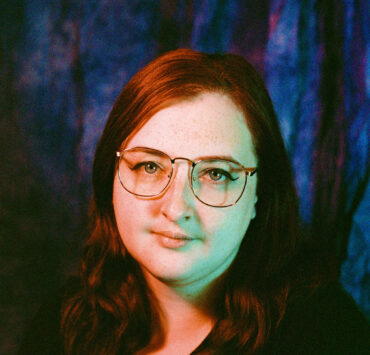 A 35mm film portrait of nat raum, a white agender person with medium-length red hair, freckles, and wire-rim glasses. A pink and blue lighting scheme washes over the author's face. Photo credit Missy Malouff.