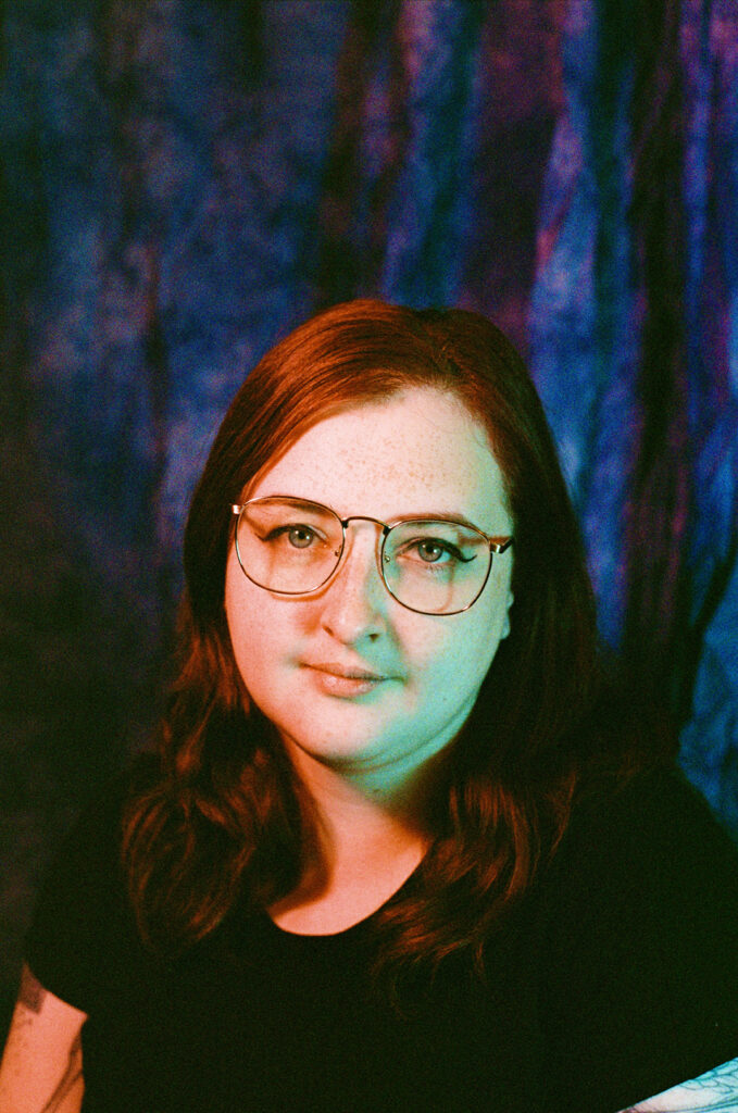 A 35mm film portrait of nat raum, a white agender person with medium-length red hair, freckles, and wire-rim glasses. A pink and blue lighting scheme washes over the author's face. Photo credit Missy Malouff.