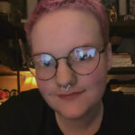 A white man with a pink buzzcut, round glasses, and a star nose ring. He is looking into the camera and smiling a little.