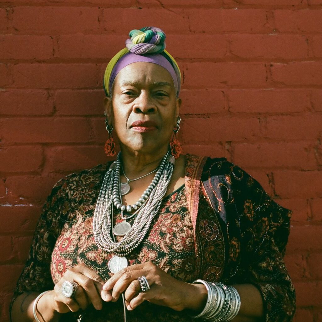 Photo of Risë Kevalshar Collins, a tall black woman whose American ancestry precedes the Civil War. She is standing before a red brick wall wearing colorful cotton headscarf, Native American silver jewelry, a Peruvian black and maroon printed dress and a Southeast Indian brown and maroon beaded shawl.