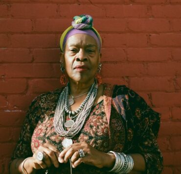 Photo of Risë Kevalshar Collins, a tall black woman whose American ancestry precedes the Civil War. She is standing before a red brick wall wearing colorful cotton headscarf, Native American silver jewelry, a Peruvian black and maroon printed dress and a Southeast Indian brown and maroon beaded shawl.