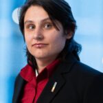 A photograph of Binx Perino, a nonbinary person with dark hair wearing a red shirt and black jacket.
