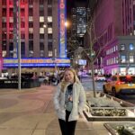 A photograph of Bree Eaton, a white woman with blond hair in a blue jacket standing outside of Radio City Music Hall in New York