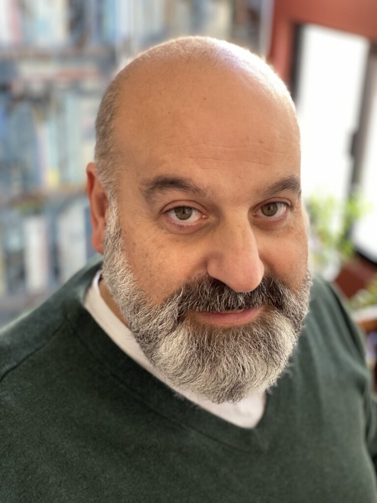 Shahé Mankerian seated in front of sagging bookshelves in his living room during the pandemic, photographed by his wife, Yvette Mankerian, shortly after learning that his first poetry collection would be published.