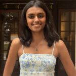 An Indian woman with shoulder-length black hair poses in front of a doorway. She is wearing a white dress with blue flowers and a green pendant necklace. She is smiling at the camera.