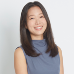 Melanie Hyo-In Han is smiling at the camera against a light grey background. She has shoulder-length dark brown hair and is wearing a slate-blue top.