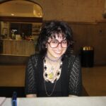 A white person with choppy bangs, black hair, and a black top sits at a table and smiles at the camera. She is wearing several necklaces and large dark red glasses.