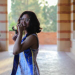 Photo of a Southeast Asian woman with glasses smiling as she looks at the camera while completing a spin, her hand hiding part of her face. She is wearing a white dress with blue flowers and a blue graduation sash with gold trim that reads, “Class of 2025” on one side and “UCLA” on the other. In the background is a brick arch that shows trees in the distance.