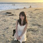 Corina Yi, an Asian American woman, sitting by the beach during sunset on her 21st birthday. She smiles at the camera in a white dress while the waves crash softly behind her.