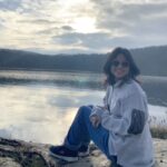 An Asian person, smiling while sitting on a rock, angled to the side, with a lake in the background. They have short black hair, light skin, and are wearing a sweater, jeans, and sunglasses.