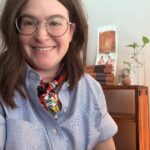 A nonbinary person with shoulder-length brown hair, big, round glasses, and dressed in a blue-striped collared shirt and colorful handkerchief is sitting and smiling for a professional headshot in front of a book shelf with art, books, and plants.