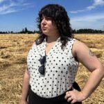 Jessica Ballen stands in a hayfield under a blue sky with her hand on her hip as she looks off into the distance. She is wearing a sleeveless white-and-black polka-dot blouse and a black skirt. Her sunglasses hang from her shirt.