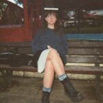 Lia Ortner, a white woman with long brown hair, sits on a wooden bench facing the camera. She is wearing a blue sweater over a dress and big black boots. She wears glasses and balances a book on her head.