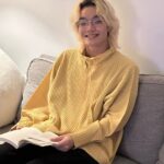 Yuanhao, an East Asian with light skin and shoulder-length blond hair, sits on a gray couch and smiles at the camera. They are wearing silver half-rim glasses, a light yellow pleated top, and black pants. An open book rests on their lap.