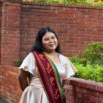 A brown Filipino woman with her hands behind her back, smiling in front of a brick-walled building. She has black hair and is wearing a gold necklace, and an ivory-colored dress with a green and maroon Sablay (or hand-woven sash) that has Indigenous geometric patterns and Baybayin characters, the official academic costume of the University of the Philippines.