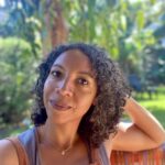 A headshot of writer Lisbeth White. She is looking directly toward the camera with a warm, small style, leaning her head back on one hand. She has medium brown skin, brown eyes, and dark curly shoulder-length hair, and is wearing a sleeveless silver tank top under brown bib-overalls. She is sitting on a colorfully woven chair with green foliage blurred behind her.