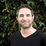 Professional author headshot of a man wearing a black shirt standing in front of a background of green ivy